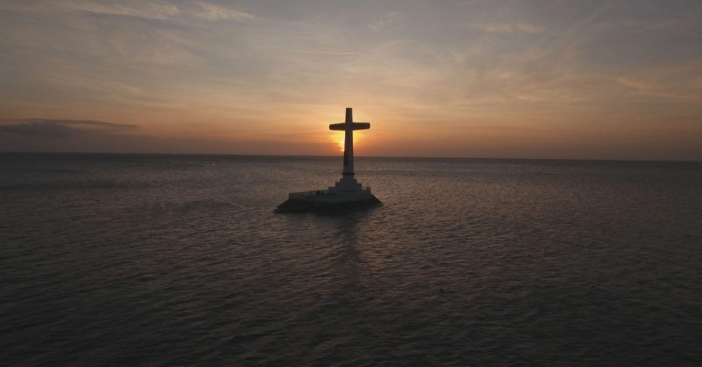 a large cross stands on a platform in the sea, silhouetted against an orange sunset—one of mindanao’s must visit spots.