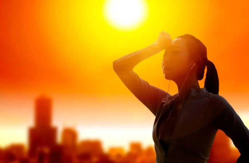 a woman in workout clothes wipes her forehead, practicing summer safety outdoors at sunset with city buildings behind her.