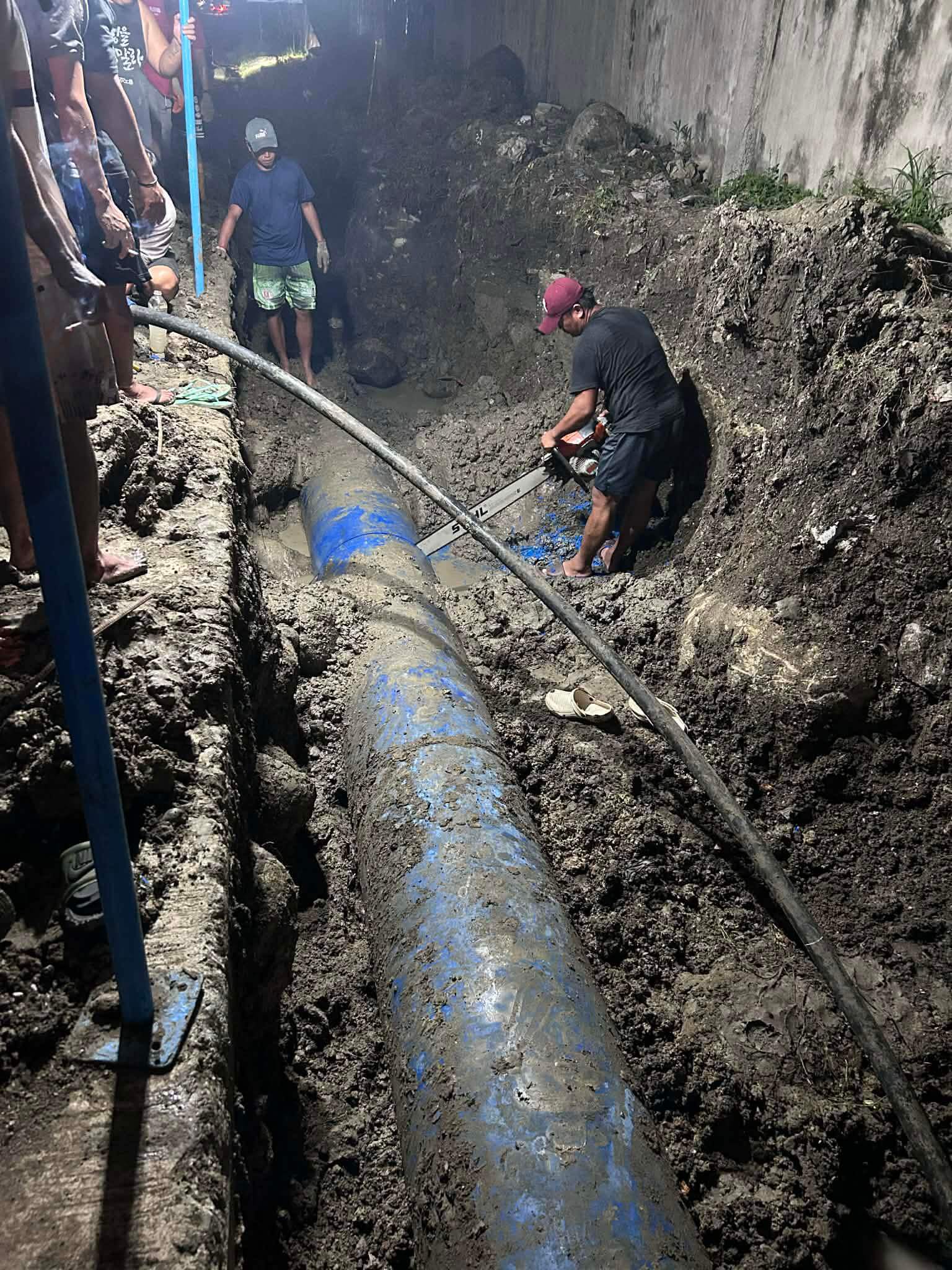 workers repair a large blue pipe in a muddy trench at night, using tools and surrounded by soil.