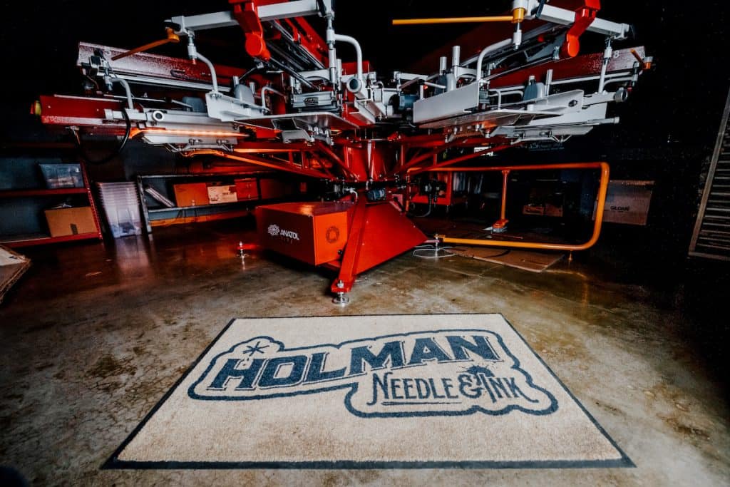 Máquina de serigrafía profesional junto a una alfombra color canela con el texto negro "Holman Needle & Ink".