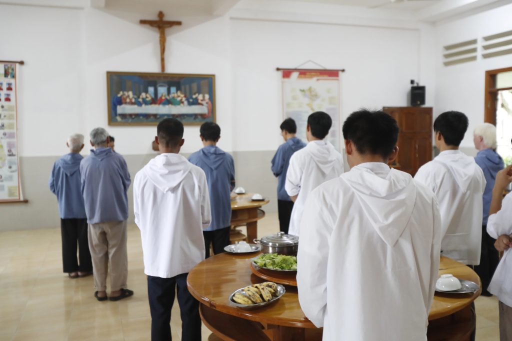Instituto Internacional de Teología a Distancia – Un grupo de personas, en su mayoría con túnicas blancas o azules, se reúne en torno a mesas con alimentos en un aula luminosa durante los Cursos para Centros. Al fondo, se aprecian un crucifijo y una pintura de la Última Cena. – formación teológica online