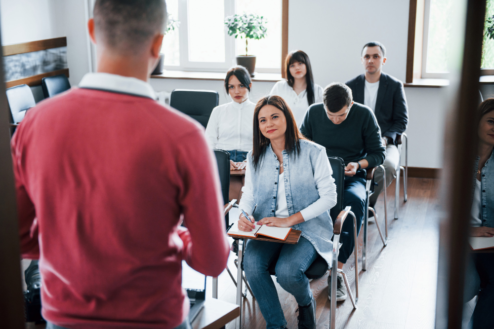Un grupo de adultos se encuentra en un aula, atento a un docente que expone; algunos toman notas. El ambiente es luminoso y ordenado.