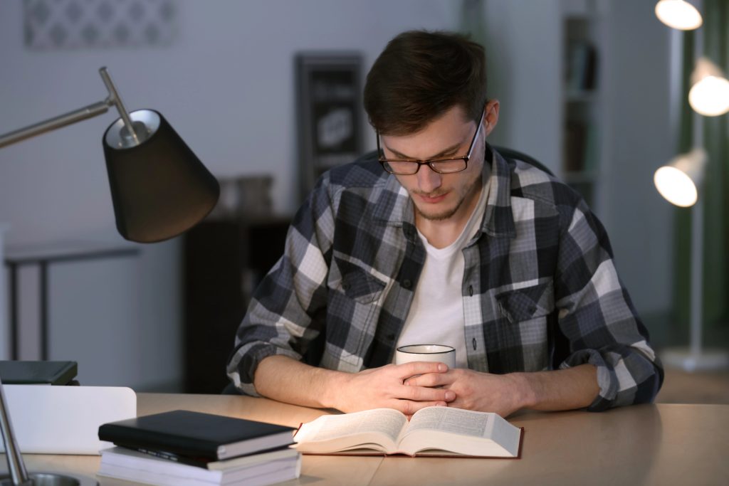 Instituto Internacional de Teología a Distancia – Una persona con gafas y camisa de cuadros lee un libro abierto sobre teología para laicos en un escritorio con lámpara, papeles y libreta. – formación teológica online