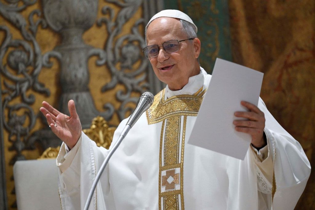 Instituto Internacional de Teología a Distancia – El Papa León XIV: Un hombre mayor, con vestiduras litúrgicas blancas y doradas, gafas y solideo blanco, expone un documento ante un micrófono en recinto decorado. – formación teológica online