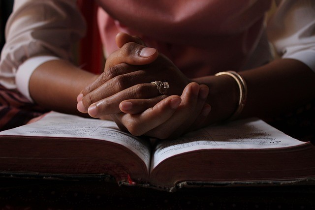Una persona con las manos juntas, apoyadas sobre una Biblia abierta, reflexiona en silencio: un símbolo de fe y dedicación a la formación bíblica. El anillo y la pulsera aportan un sutil detalle a la escena, bañada por una luz tenue.