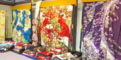 色とりどりの着物と反物が所狭しと並べられた部屋。着物には、鮮やかな青、赤、紫の色合いで、花や自然をテーマにした繊細な模様が描かれています。展示された着物の下には、様々な色の反物が重ねられています。.