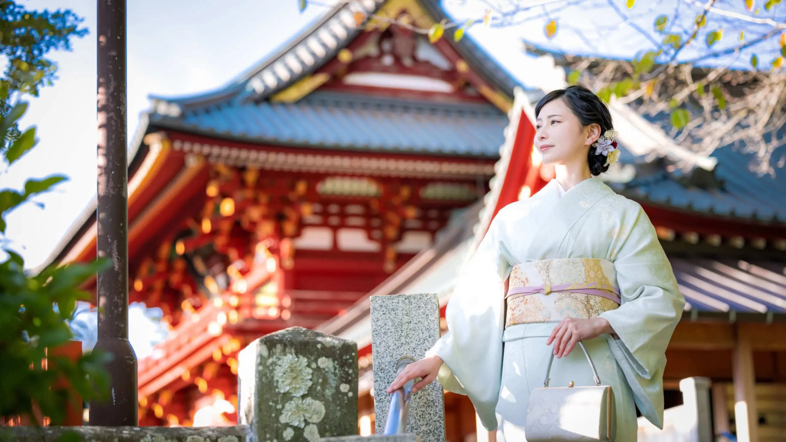 白い着物を着た女性が、伝統的な日本の寺院の前に立っています。彼女は小さなベージュのハンドバッグを持ち、髪には花を飾っています。背景には、赤と白の精巧な装飾が施された寺院が部分的に見えています。明るい日差しが、この光景を照らしています。.