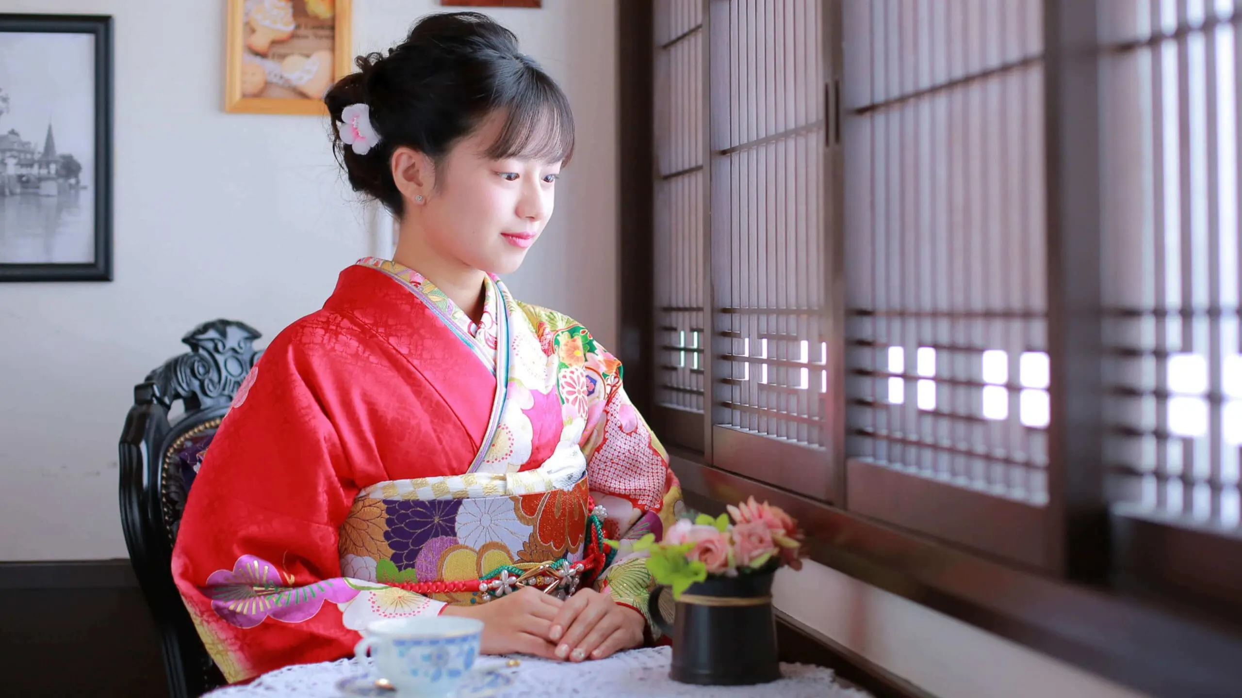 赤い花柄の着物を着た女性が、レースのテーブルクロスがかけられたテーブルに座り、木製の雨戸のついた窓の外を眺めている。テーブルの上には小さな生け花とティーカップが置かれている。.