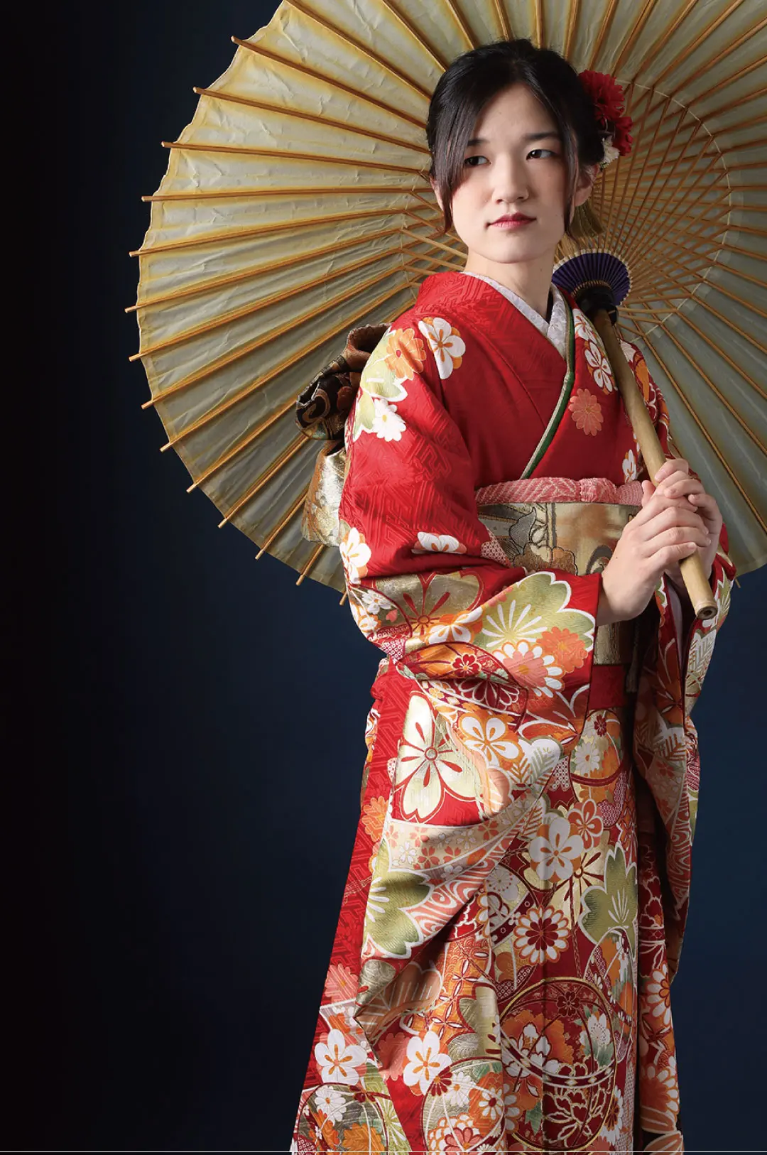 赤い花柄の着物を着た人物が、大きなベージュ色の日傘を差しています。髪には花を飾り、暗い背景に立って横を見つめています。.