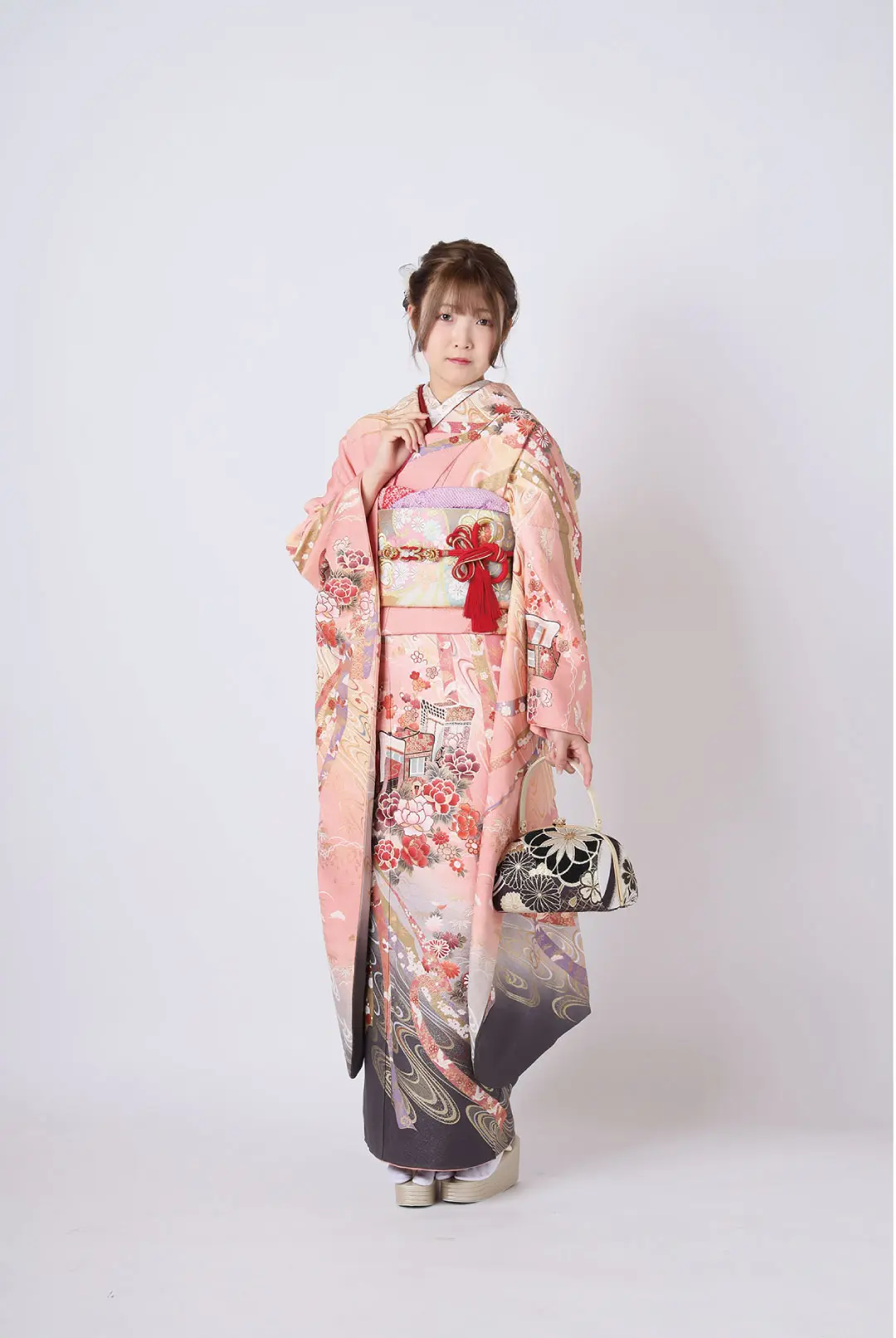 シンプルな背景に、令和6年成人式の伝統的なピンク色の着物を着た人物が立っています。着物は複雑な花柄があしらわれた伝統的な着物です。小さな柄のハンドバッグを持ち、白い草履を履き、髪はきちんとセットされ、装飾品で飾られています。.