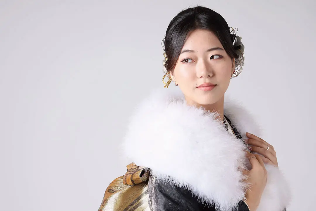 ふわふわの白い襟と華やかな髪飾りをつけた、落ち着いた表情の伝統的な令和6年成人式の着物を着た女性が、金色の模様のバッグを肩にかけて、無地の背景を横目で見ています。.