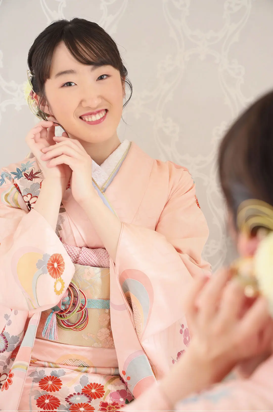 微笑みながら、花柄があしらわれたパステルピンクの着物を着た人が鏡を見て、複雑な模様の背景を背景にイヤリングを直している。.