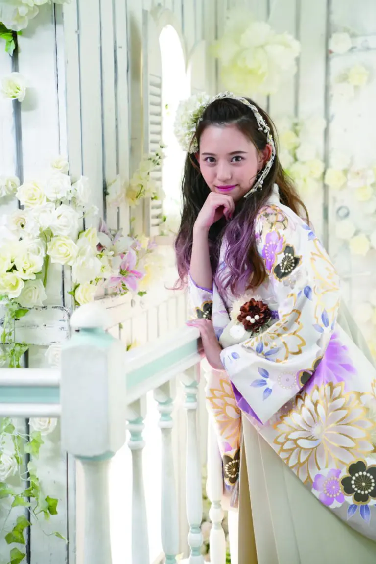 白い木製パネルとたくさんの花々を背景に、長い髪を部分的に紫に染めた花柄の着物「レンタル袴 01」を着た女性が白い手すりに寄りかかりながら微笑んでいます。.