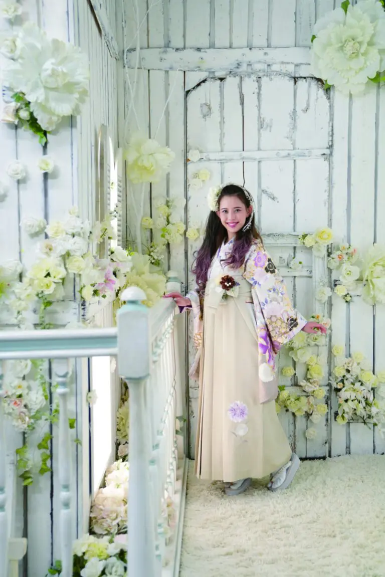 レンタル袴 01 を着た女性が、素朴な白を基調とした部屋、古びた木目調の壁、柔らかな照明の中で、大きな白とクリーム色の花に囲まれた白い木製の階段に立っています。.