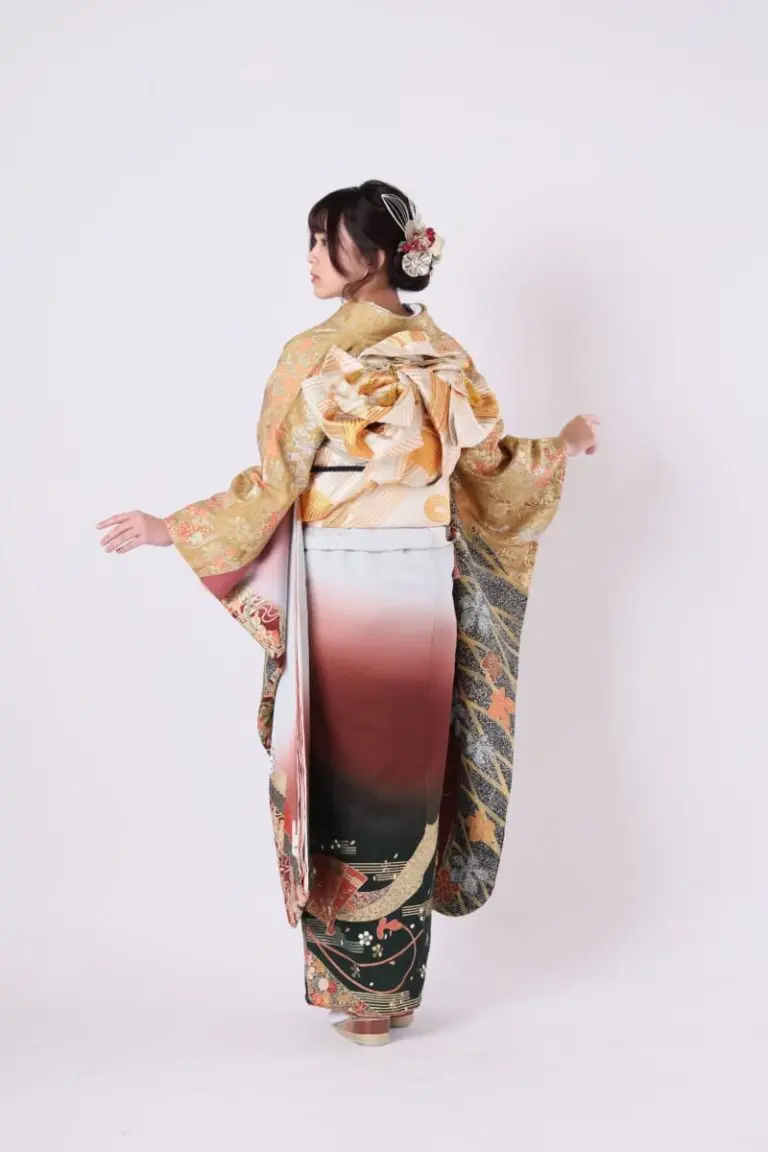 複雑な模様が描かれた日本の伝統的な着物を着た女性が、無地の白い背景に立っています。髪には花飾りが飾られ、カメラに背を向け、両腕を少し伸ばしたポーズをとっています。.