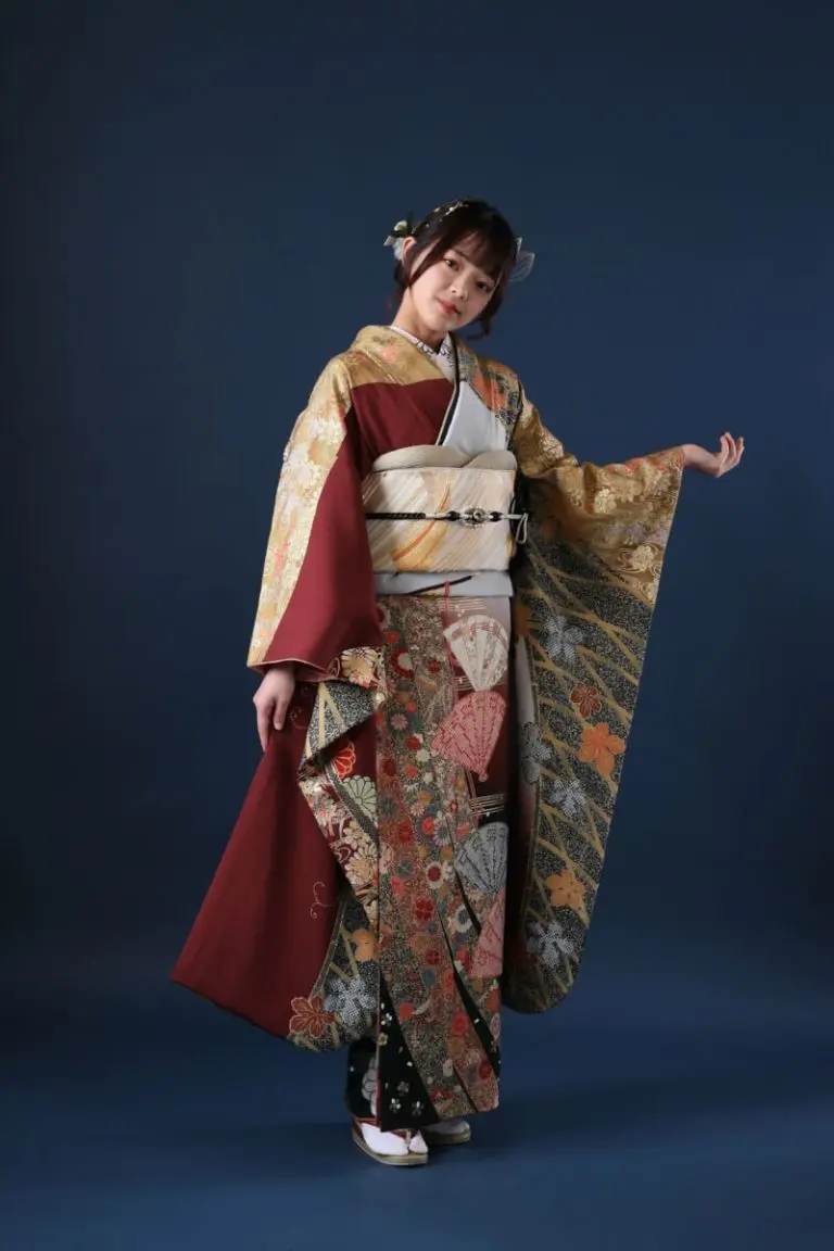 暗い背景に、赤、金、黒の複雑な模様が描かれた日本の伝統的な着物を着た人物が立っています。髪には花飾りが付けられ、着物の袖は少し横に広げられています。.