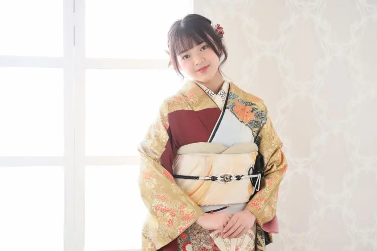 室内に、花柄の着物を着た女性が立っています。彼女は少し横に寄り、両手を前に出しています。背景には模様のある壁と、左側に窓があります。.