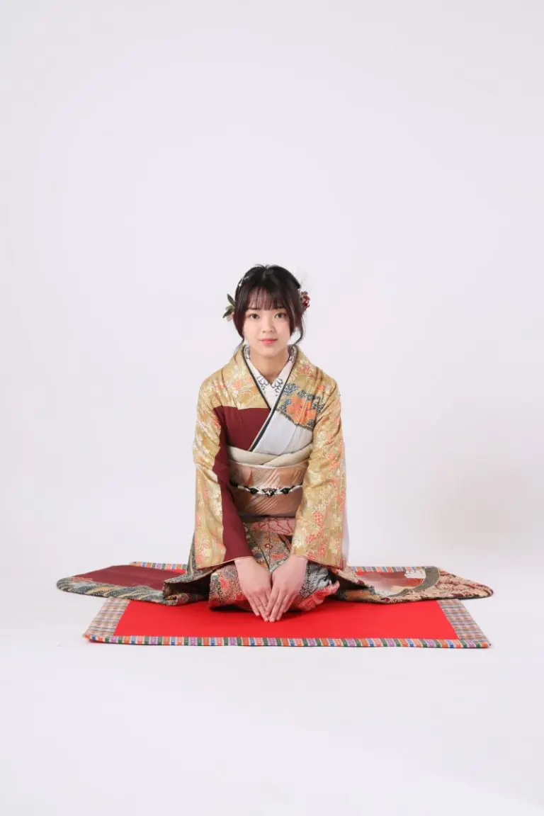 複雑な模様が描かれた日本の伝統的な着物を着た人が、無地の白い背景に敷かれた色とりどりのマットの上に座っている。彼らは正座し、両手を膝の上に置いている。.