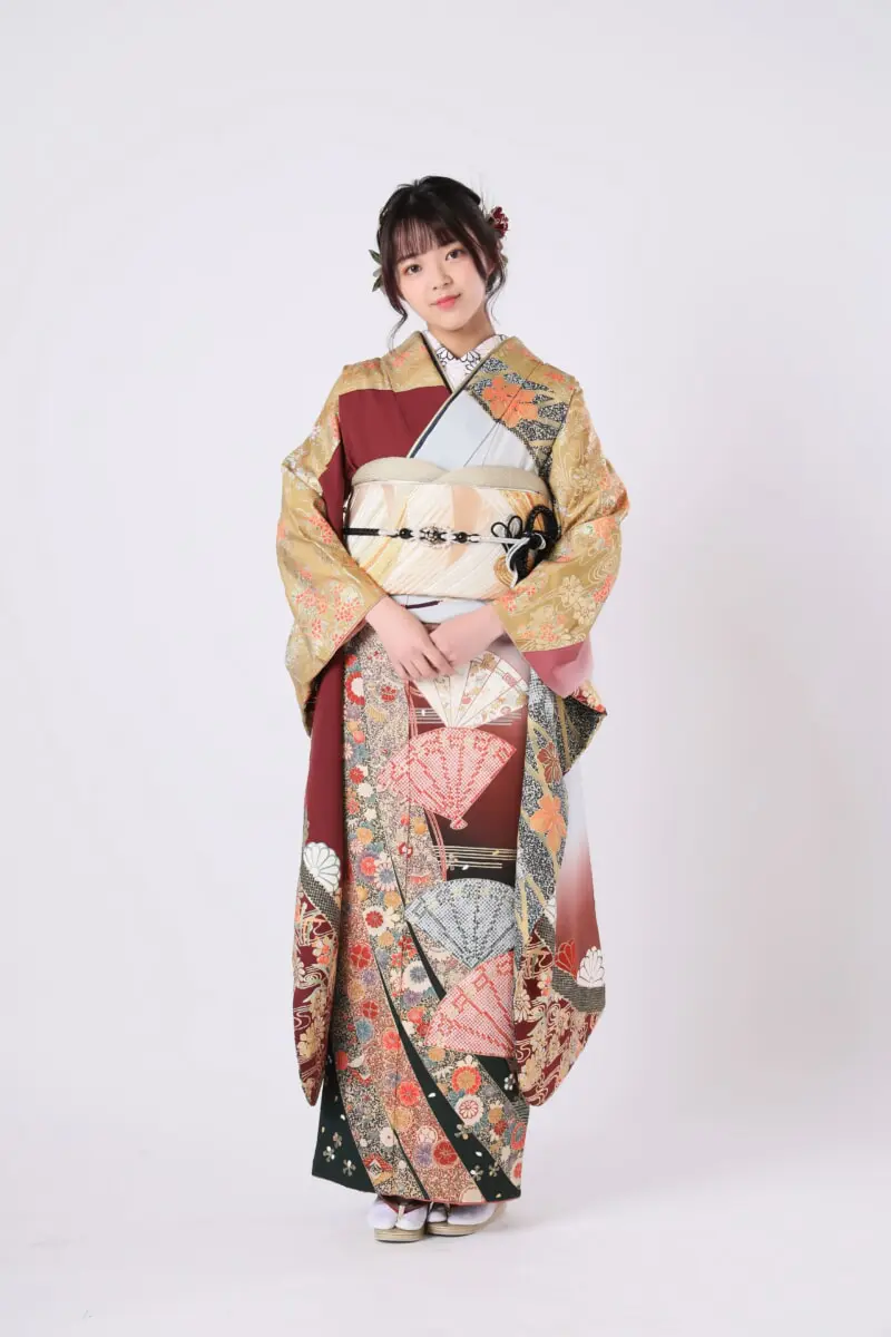 色鮮やかで複雑な模様の着物を着た人物が、無地の背景に立っています。両手を前に組んでおり、花の髪飾りをつけています。.