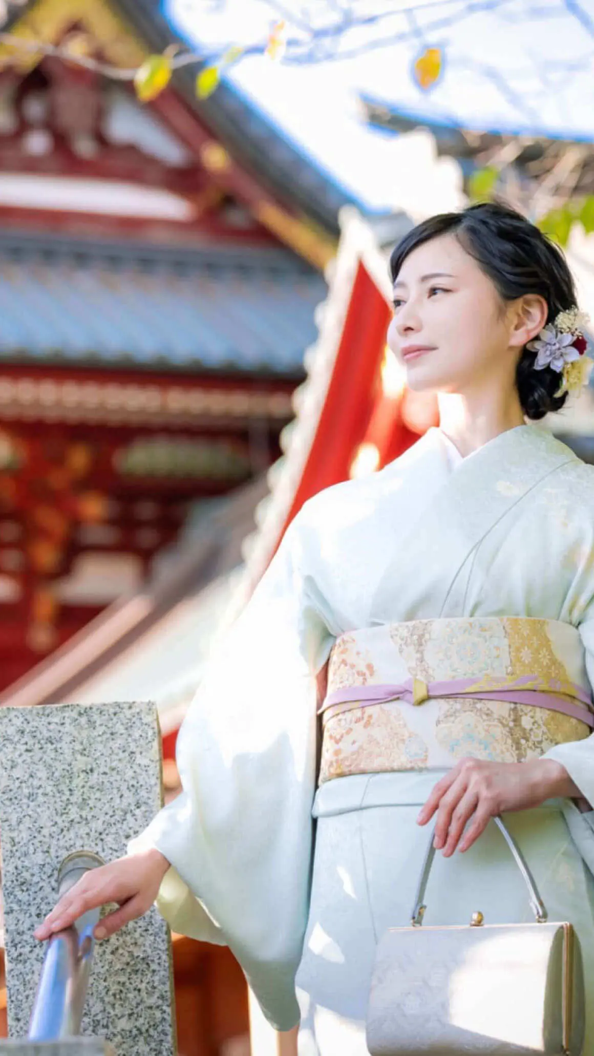 淡い色の着物を着た女性が、伝統的な日本建築の近くの屋外に立っています。彼女は小さな淡い色のハンドバッグを持ち、髪には花を飾っています。背景には建築の細部と鮮やかな色が描かれています。.