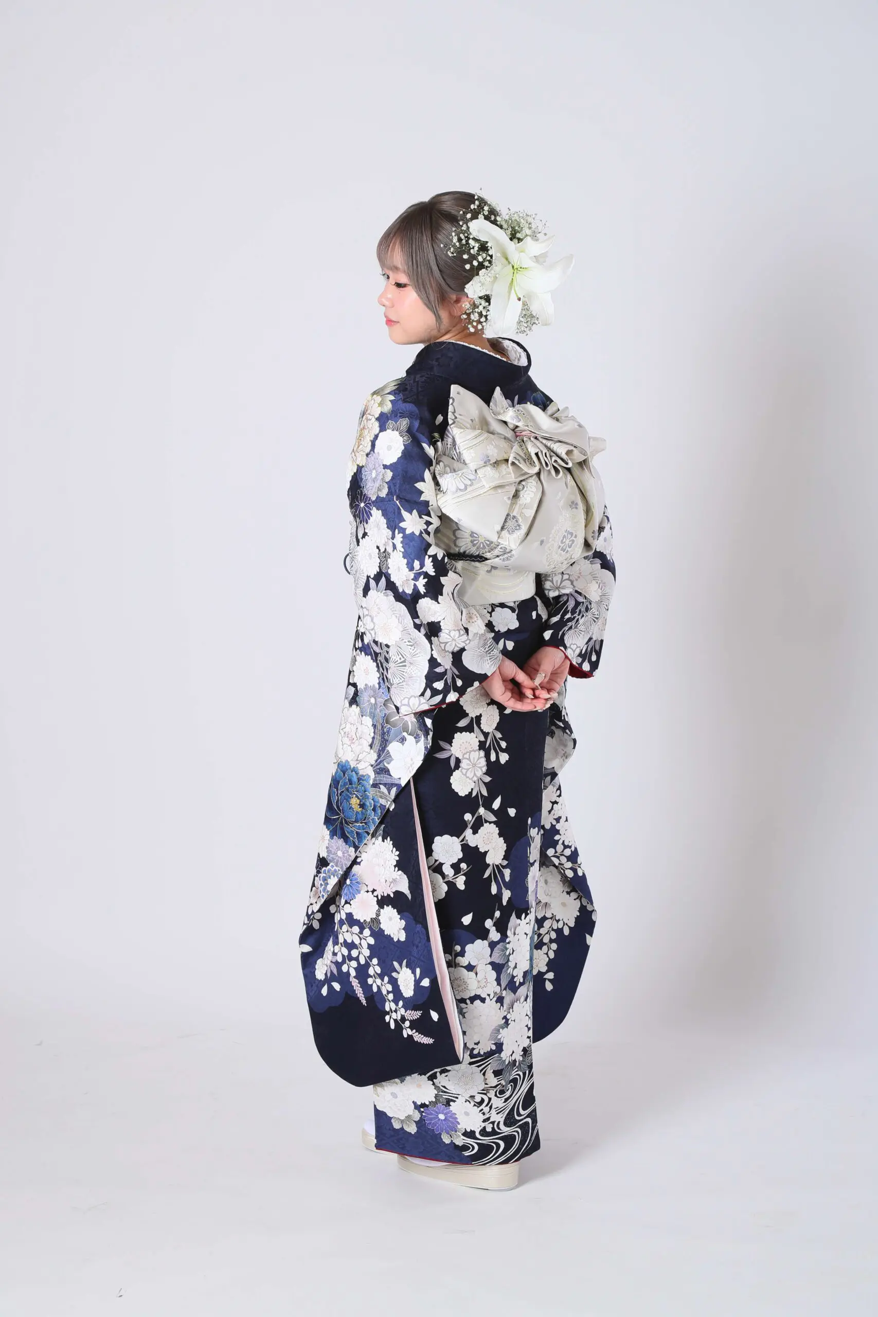 白い花柄が入った紺色の着物を着た人物が、無地の白い背景に立っています。短い髪には白い花が飾られ、着物の背中には大きな飾りのリボンが結ばれています。両手を背中に組んで、横を向いています。.