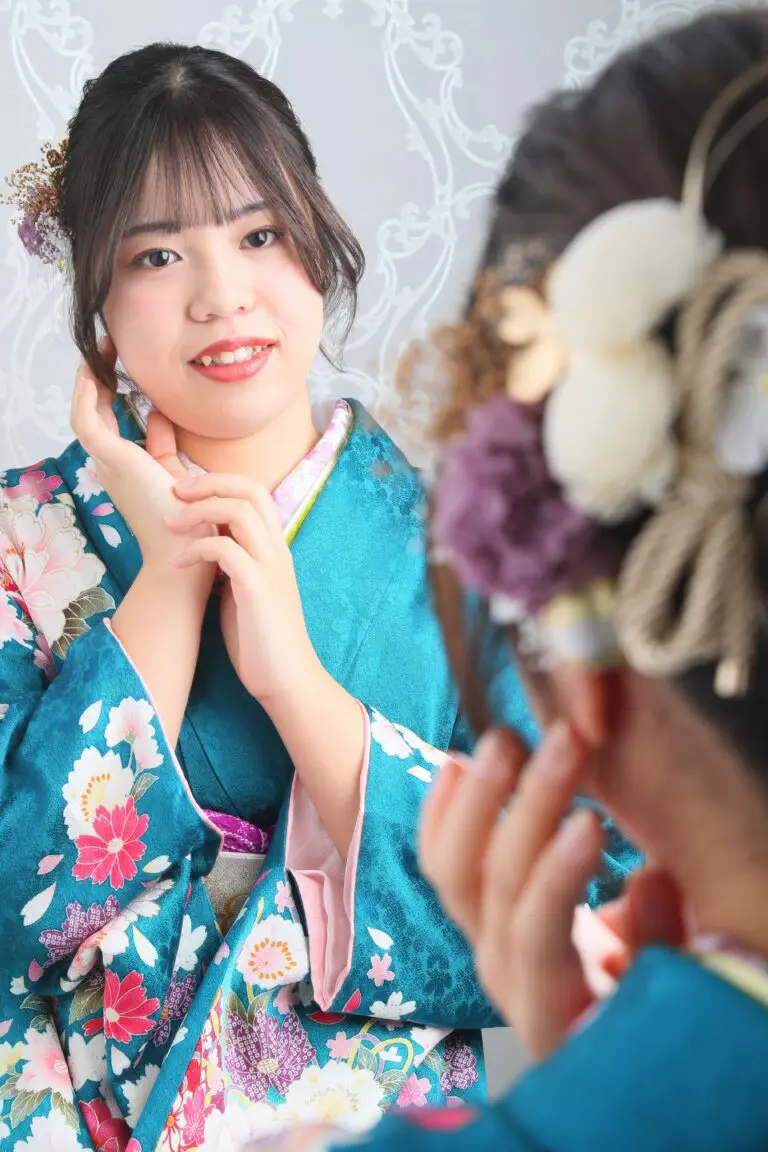 令和7年成人式、蕨市コレクションの青い花柄の着物を着た人が、模様のある背景に自分の顔を触ったり、花で飾られた髪型を整えたりしながら、鏡に映る自分の姿を眺めています。.