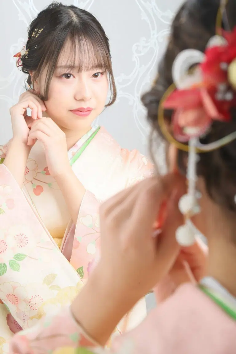 令和7年成人式、蕨市のために花柄のピンクの着物を着た彼女は、微妙な模様の白い背景に赤と白の飾りで飾られた髪を鏡で見てイヤリングを直している。.