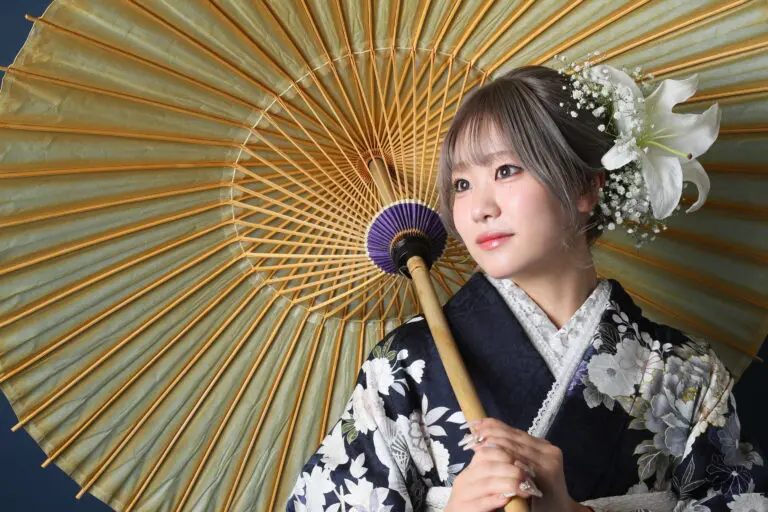 白黒の花柄の着物を着た人物が、大きな黄色の日傘を差しています。髪には白い花が飾られ、暗い背景を背景に、無表情で横を向いています。.