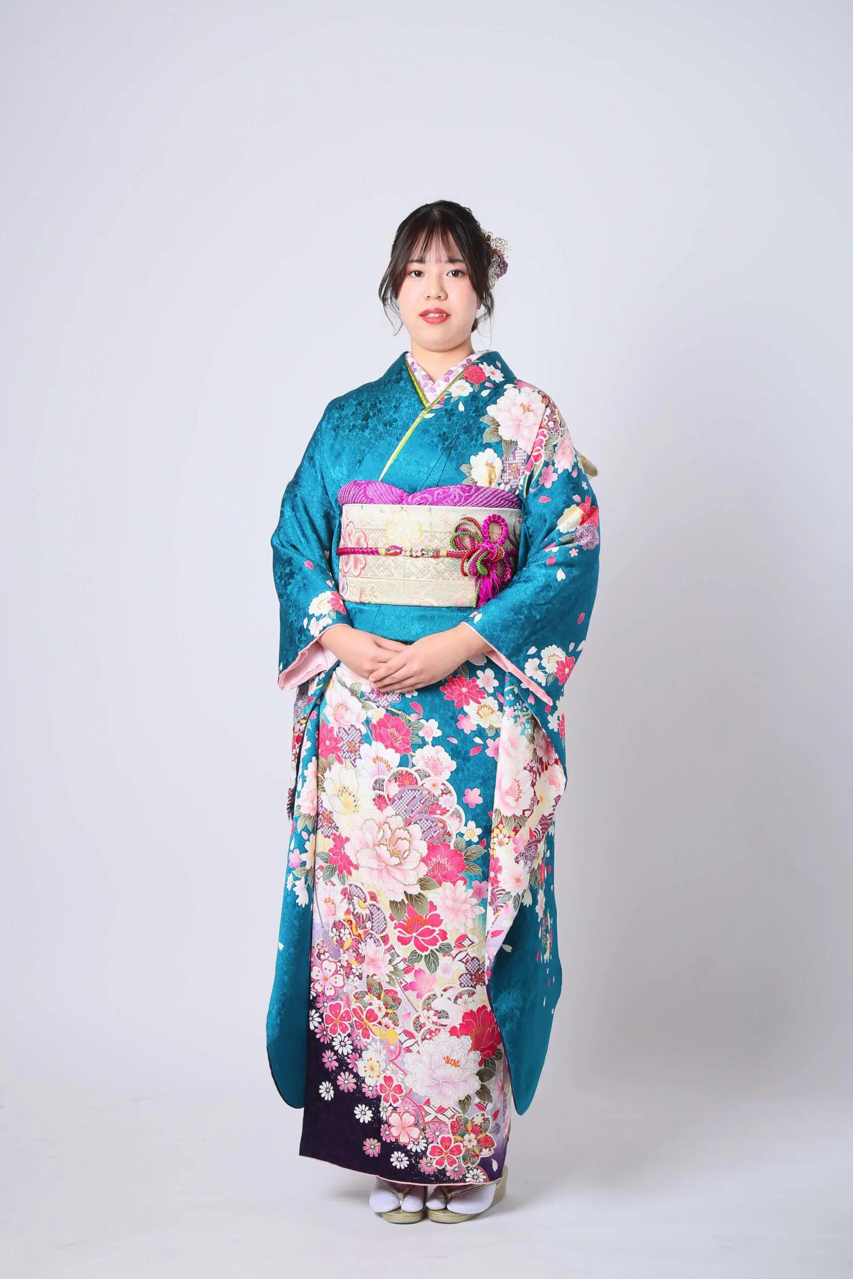 令和7年成人式、蕨市のイベントで、ピンクと白の花柄があしらわれたターコイズブルーの着物に、それに合わせた帯を締めた人物が着用しています。髪には、無地のライトグレーの背景に花飾りが施されています。.