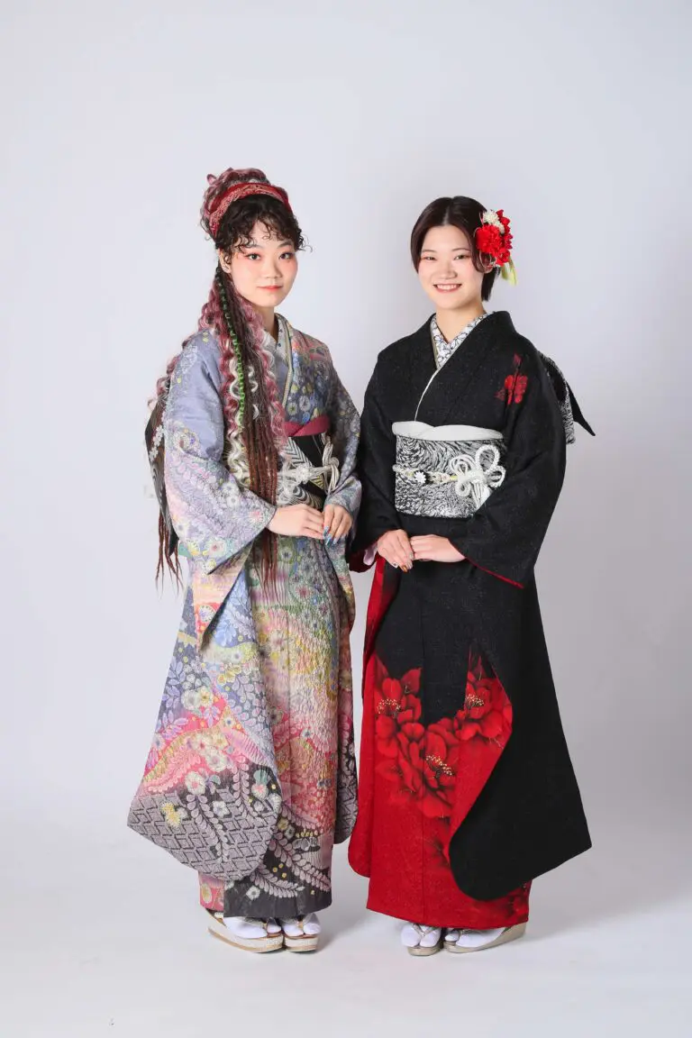 白い背景に、ヘアスタイルを整え、着物を着た二人の女性が立っています。一人は色鮮やかで繊細な模様の花柄の着物、もう一人は黒と赤の花柄がアクセントになった着物です。衣装は「令和7年成人式、戸田市」コレクションのものです。二人とも笑顔で、優雅さを体現しています。.
