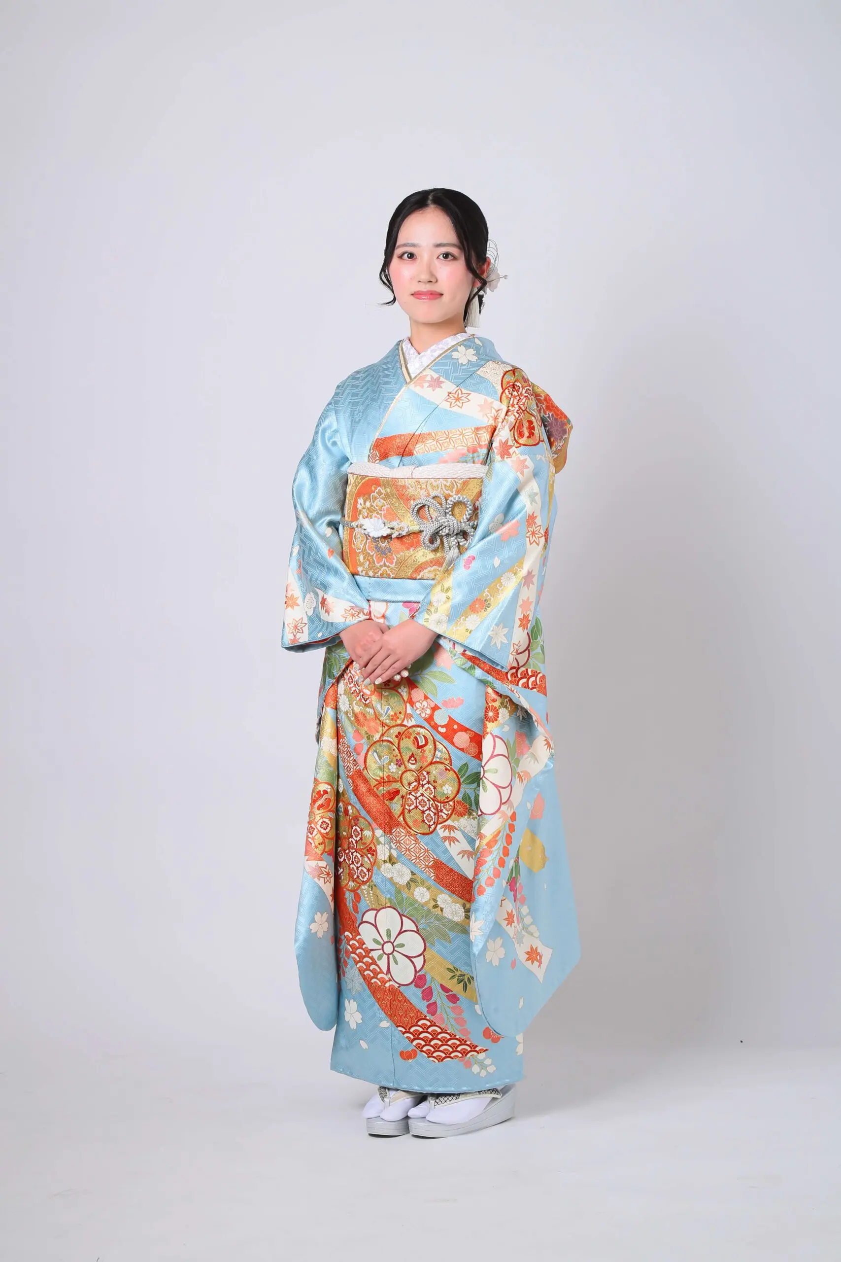 無地の背景に、色鮮やかな模様が入った伝統的な青い着物を着た人物がカメラに向かって立っています。両手を組み、白い足袋と草履を履いています。.