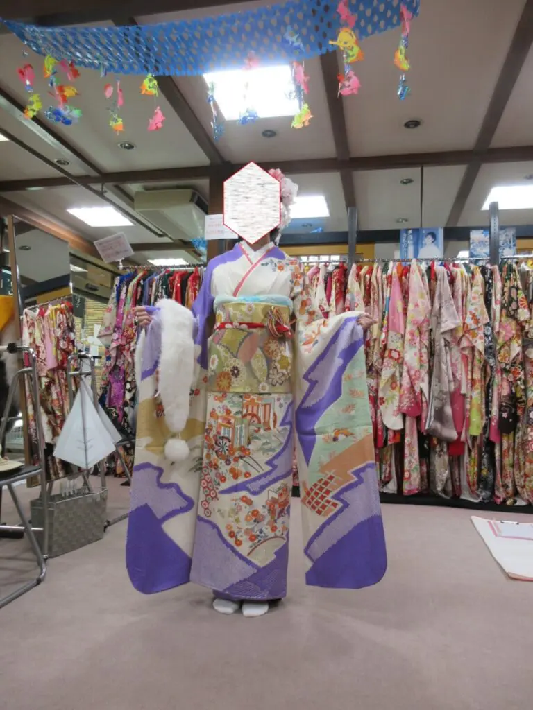 屋内の振袖展で、複雑な模様と紫の袖の着物を着た人が、色とりどりの着物が並べられた棚の前に立っている。顔は六角形の飾りで隠されており、天井からは飾りが吊り下げられている。.