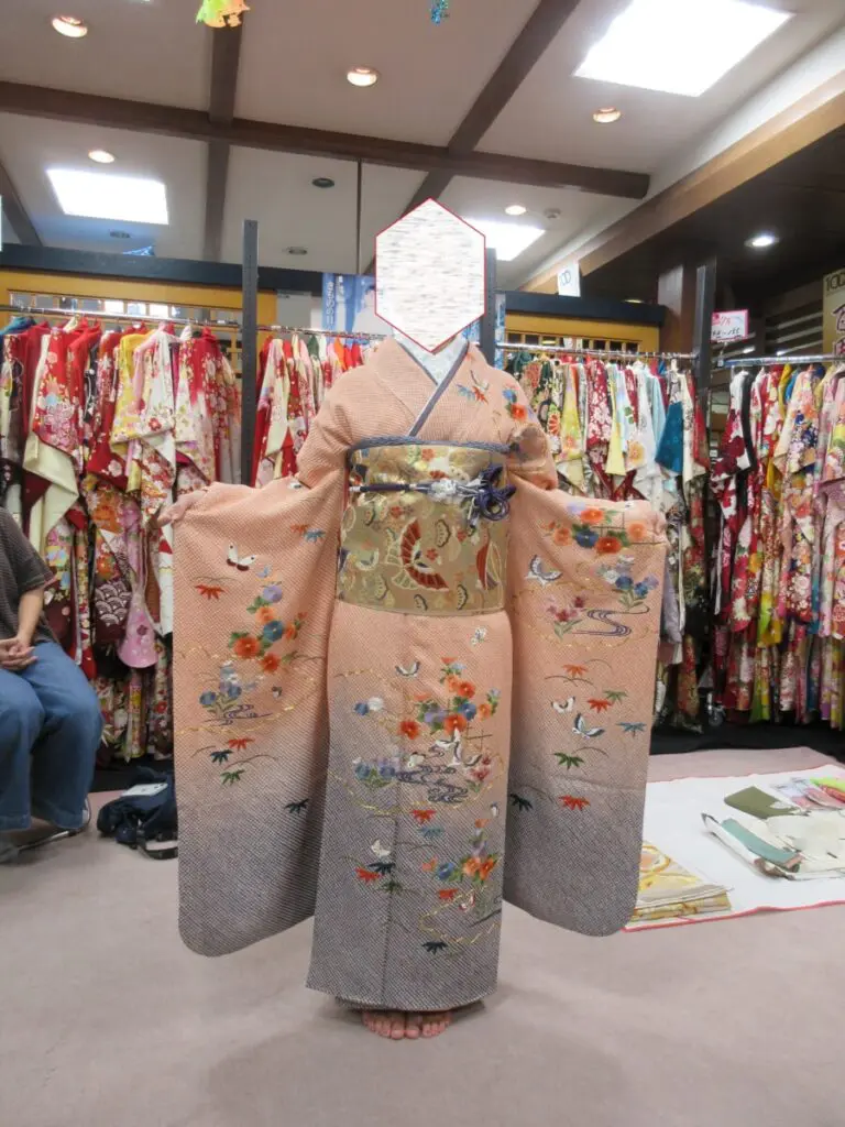 華やかな振袖展が開かれる中、伝統的な模様の着物を着た人が室内で両腕を広げて立っている。背景には色とりどりの着物がラックに並べられており、人物の顔は見えていない。.