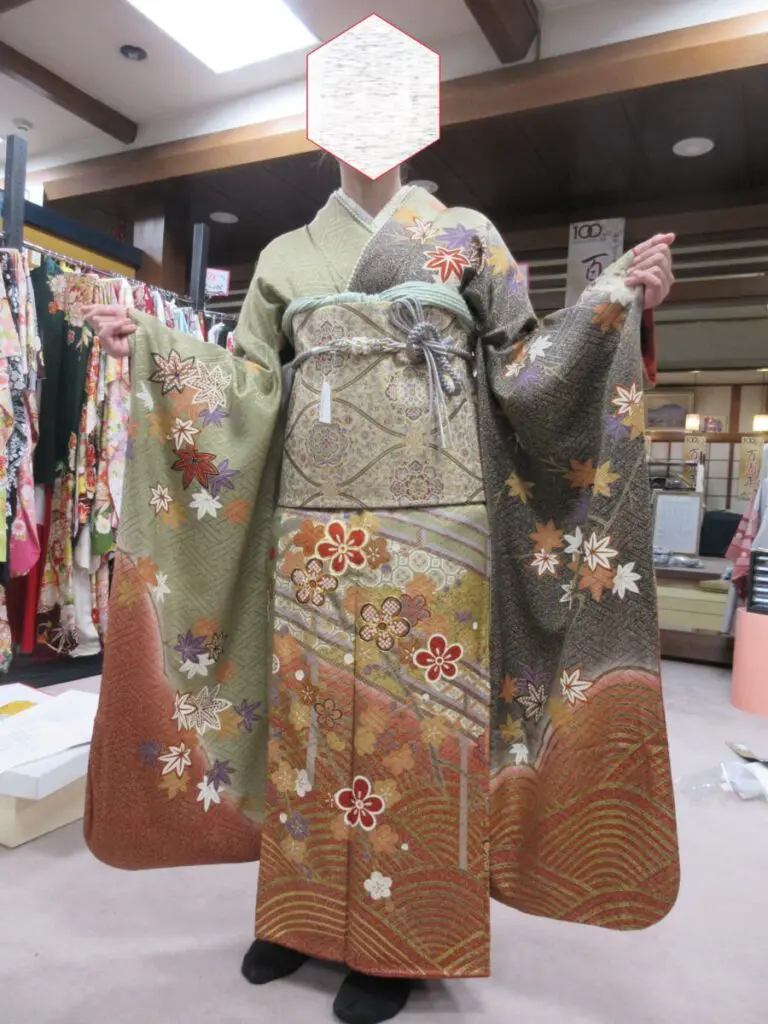 緑、茶、オレンジ色の紅葉と花柄の伝統的な着物を着た人物が室内に立っている。顔は隠されている。振袖展の背景には、他の着物が掛けられた洋服掛けが見える。.