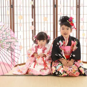 二人の幼い子供が、飾り板の前の畳の上に座っています。二人とも日本の伝統的な着物を着ていて、下の子は白とピンクの着物を、上の子は黒とピンクの着物を着ています。.