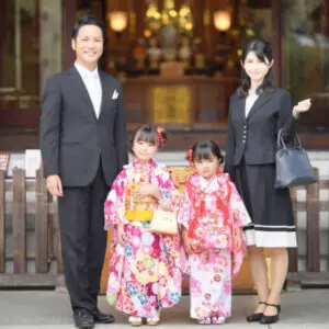 正装した男女が、色鮮やかな着物を着た二人の少女とともに、日本の伝統的な建物の外で写真撮影に応じている。.