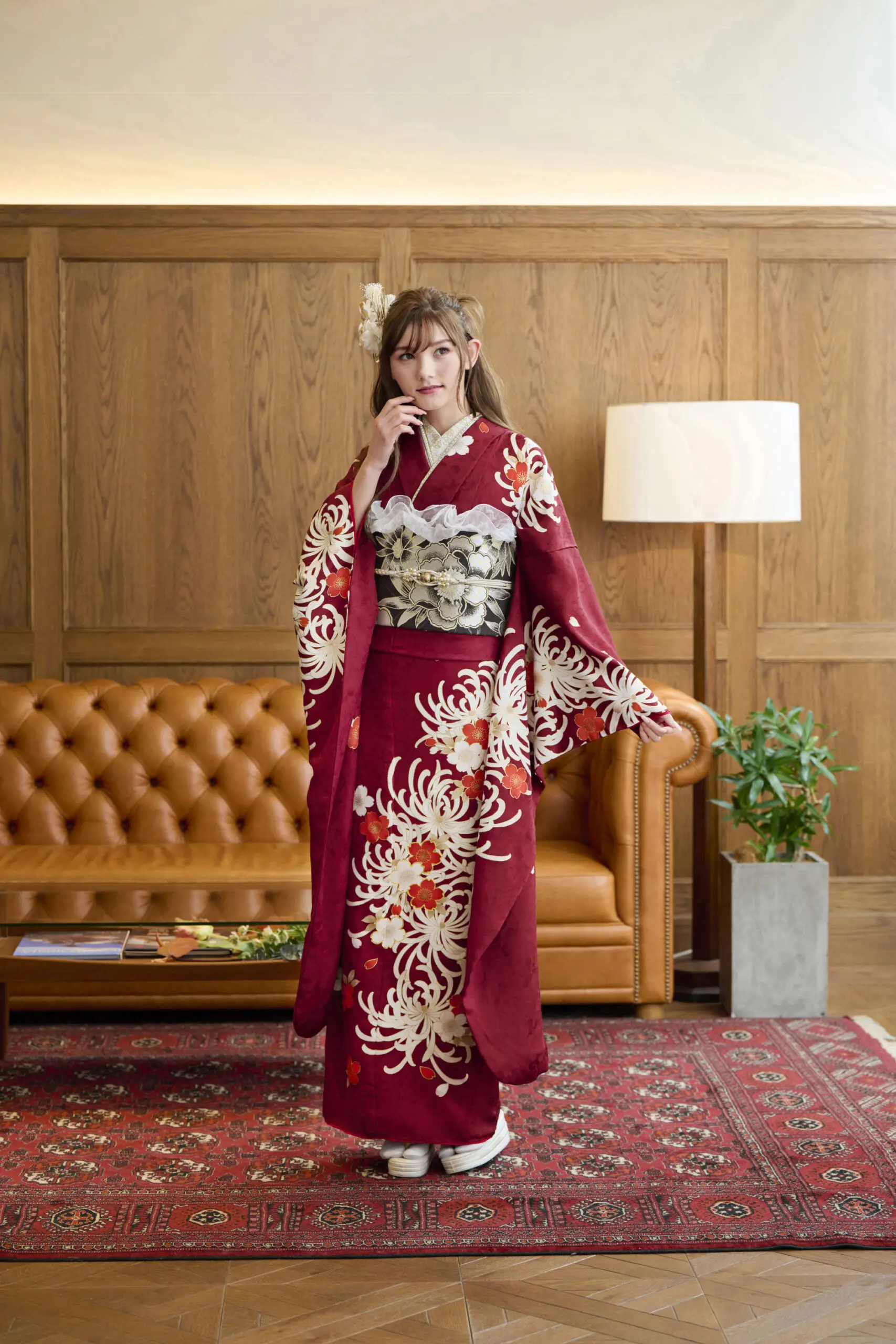 茶色の革張りのソファ、木製の羽目板、フロアランプ、鉢植えのある部屋で、M-404の赤い花柄の着物を着た人が柄物のラグの上に立っている。.