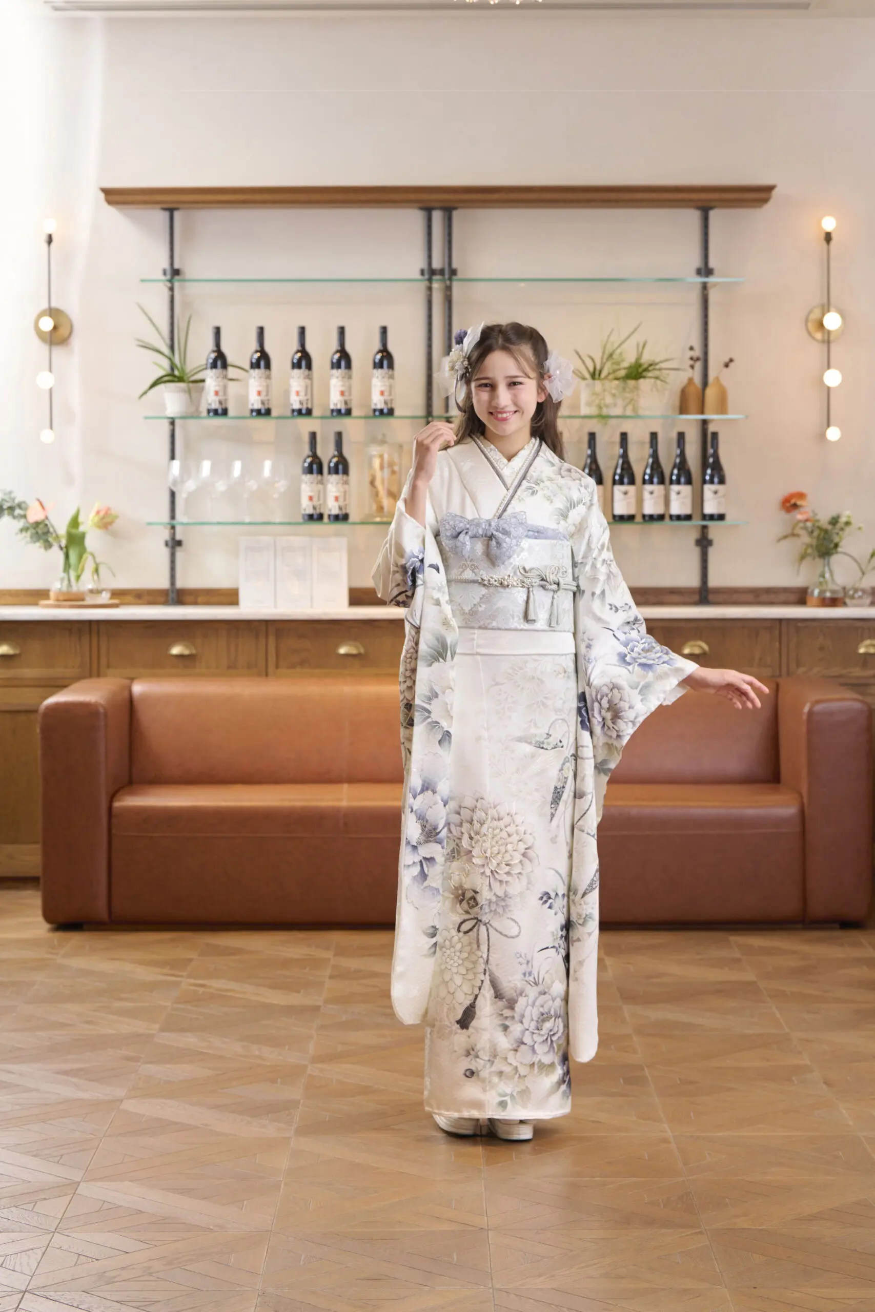 茶色のソファと棚の前で、M-410の白い花柄の着物に帯と髪飾りを合わせ、片袖を出して微笑む女性。.
