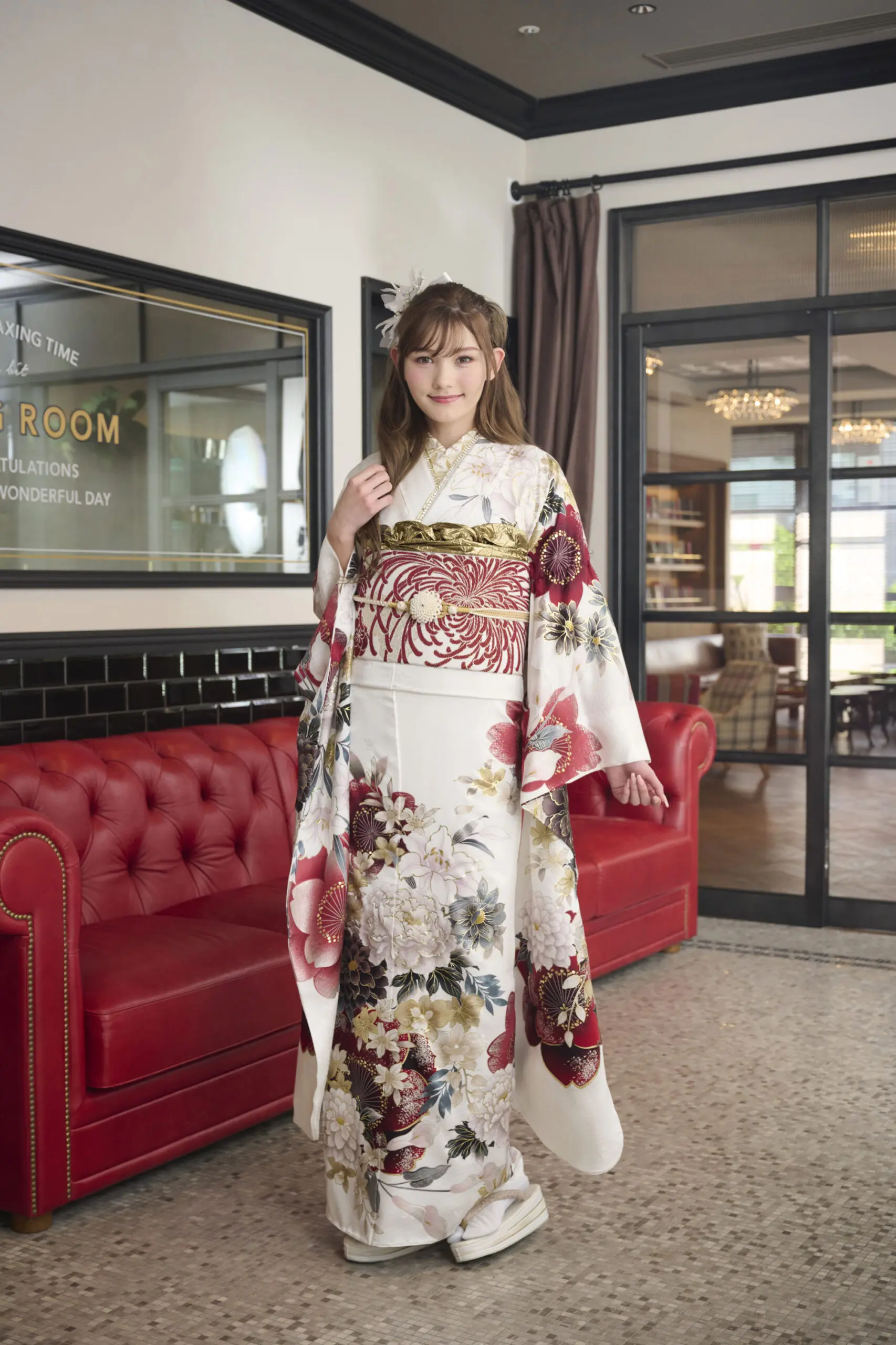 M-403-赤と金の模様が入った白い花柄の着物を着た若い女性が微笑みながら室内に立っている。片手を少し上げ、背後には赤いソファと額縁入りの鏡が置かれている。.