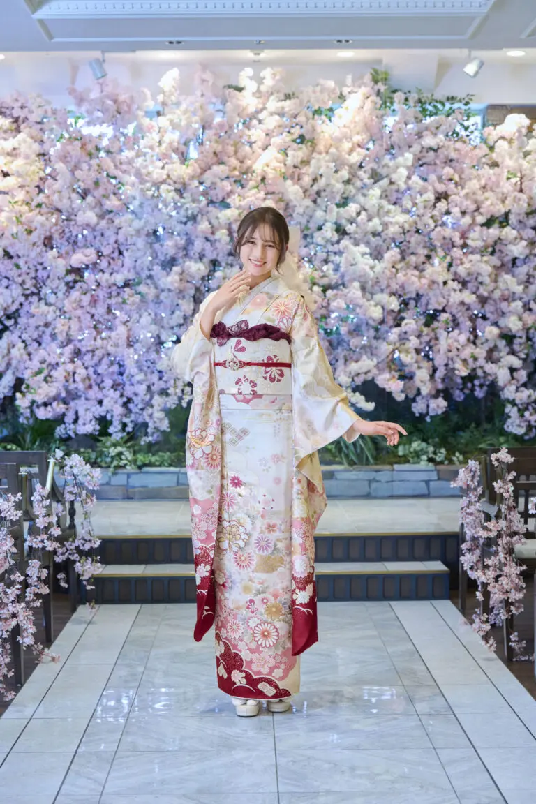 段差のあるタイル張りの小道でM-408の花柄の着物をモデルにしている女性が、ピンクと白の桜が咲き誇るエレガントな照明の前でポーズをとっている。.