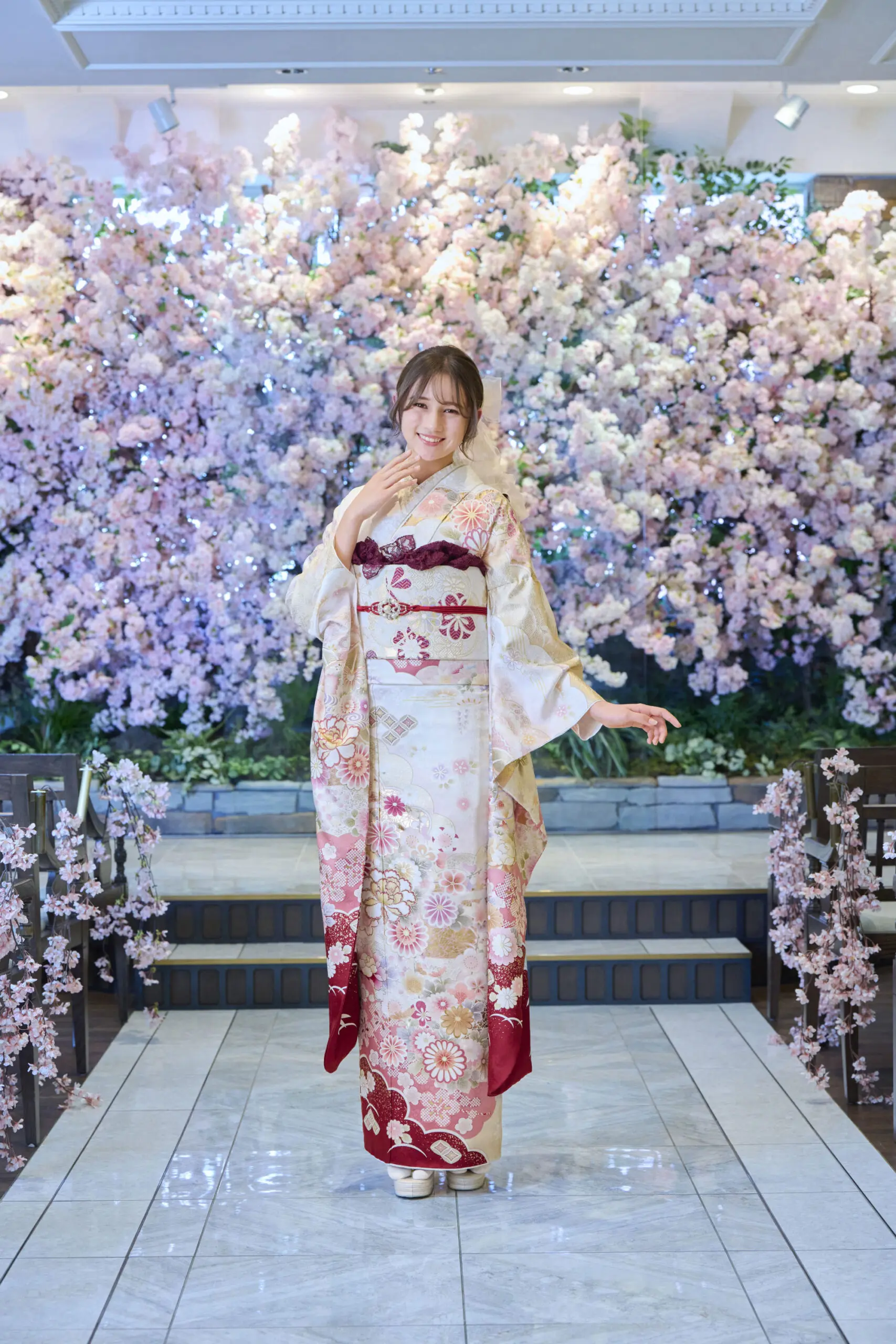段差のあるタイル張りの小道でM-408の花柄の着物をモデルにしている女性が、ピンクと白の桜が咲き誇るエレガントな照明の前でポーズをとっている。.