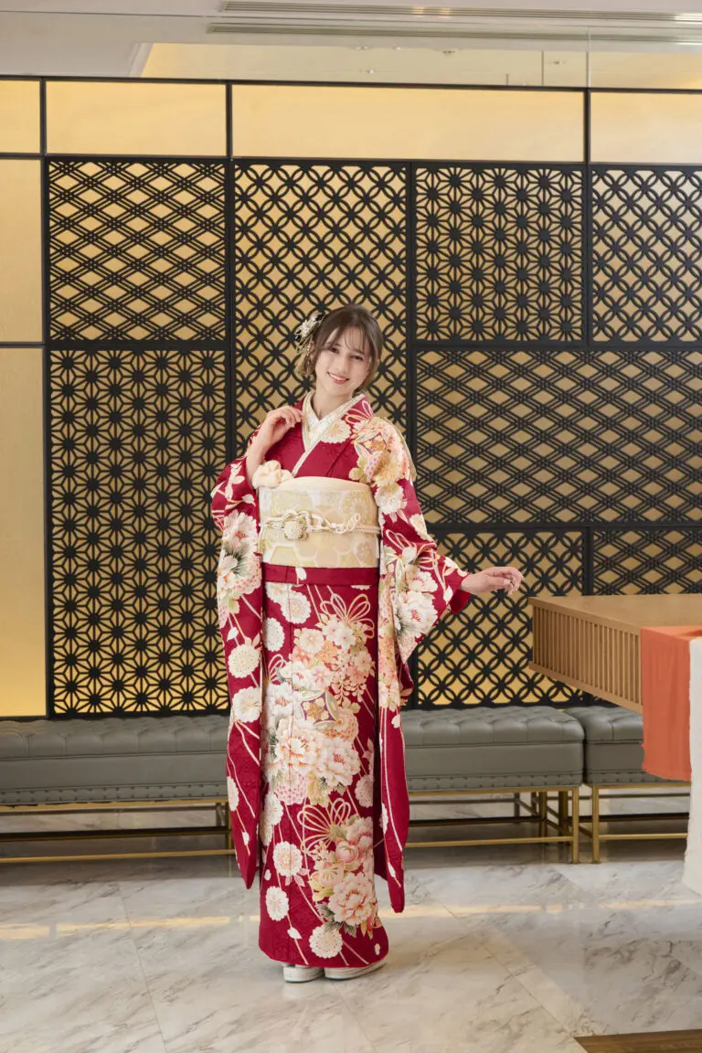 M-414の赤い花柄の着物にクリーム色の帯を締め、笑顔で片手を上げてポーズをとる人物が室内に立っている。背景は幾何学模様の装飾パネルと大理石の床。.