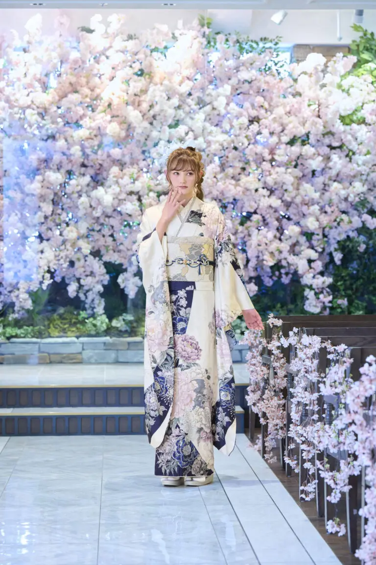 M-402柄の着物を着た人物が、屋内の磨き上げられた床の上に立っている。背景には満開の桜の木があり、階段の横に立っている。.