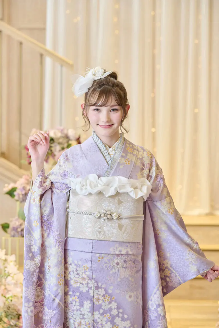 M-401の薄紫の花柄の着物に銀の帯と白いリボンをつけ、室内でカメラに向かって微笑む若い女性。薄茶色の髪に白いアクセサリー。淡い花とドレープが背景を飾る。.