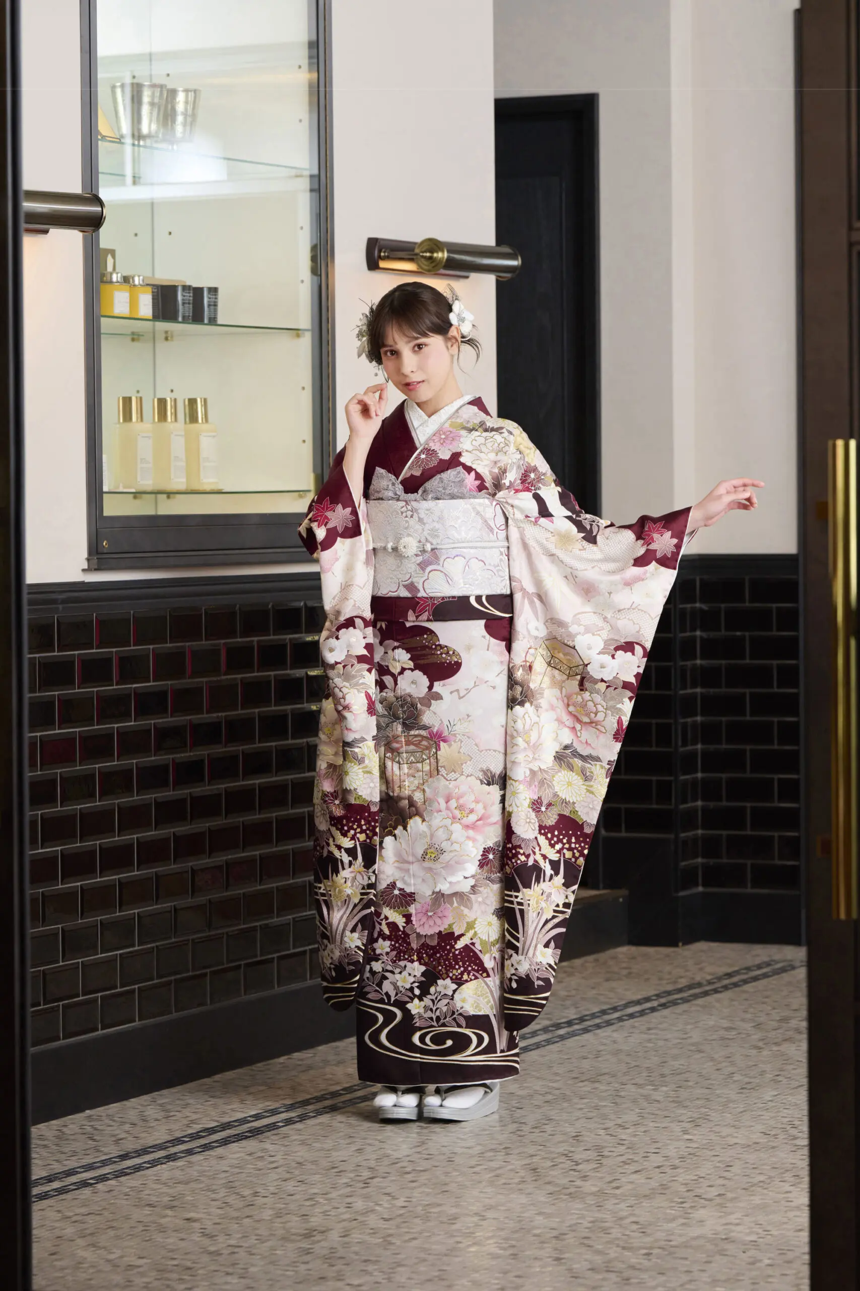白、ワインレッド、ゴールドの花柄が特徴的なM-421フォーマル着物を着て、室内でポーズをとる女性。髪飾りをつけ、片手で顔を触り、もう片方の手で着物の袖を持っている。.
