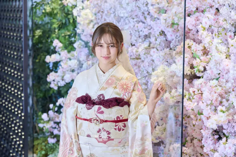 ピンクの桜と緑の壁の前に、赤と金のアクセントが入ったM-408の淡い色の着物を着た女性が、ニュートラルな表情でカメラを見つめている。.