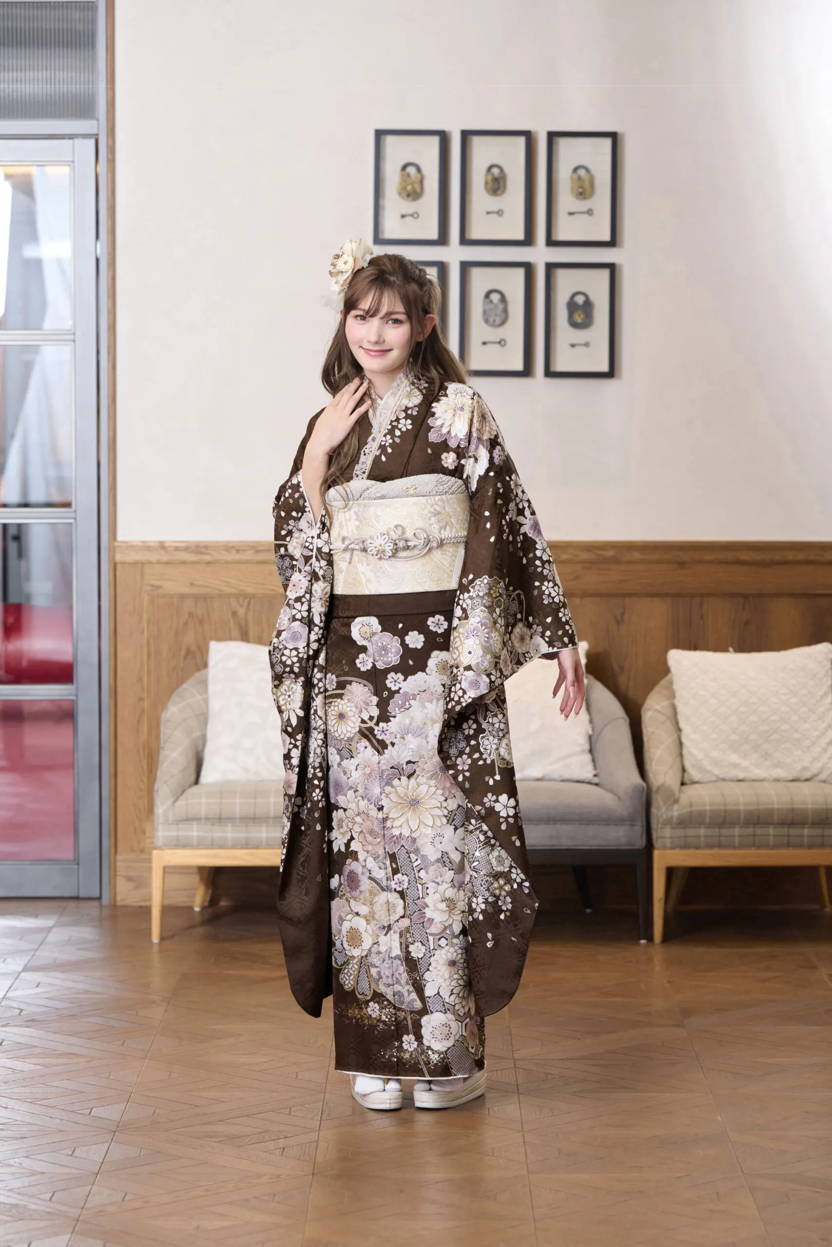 M-405の茶色の花柄の着物を着て、ベージュの帯を締めた女性が室内に立っている。髪に花を飾り、2つのソファの前でポーズをとり、後ろの壁には額装された肖像画が飾られている。.