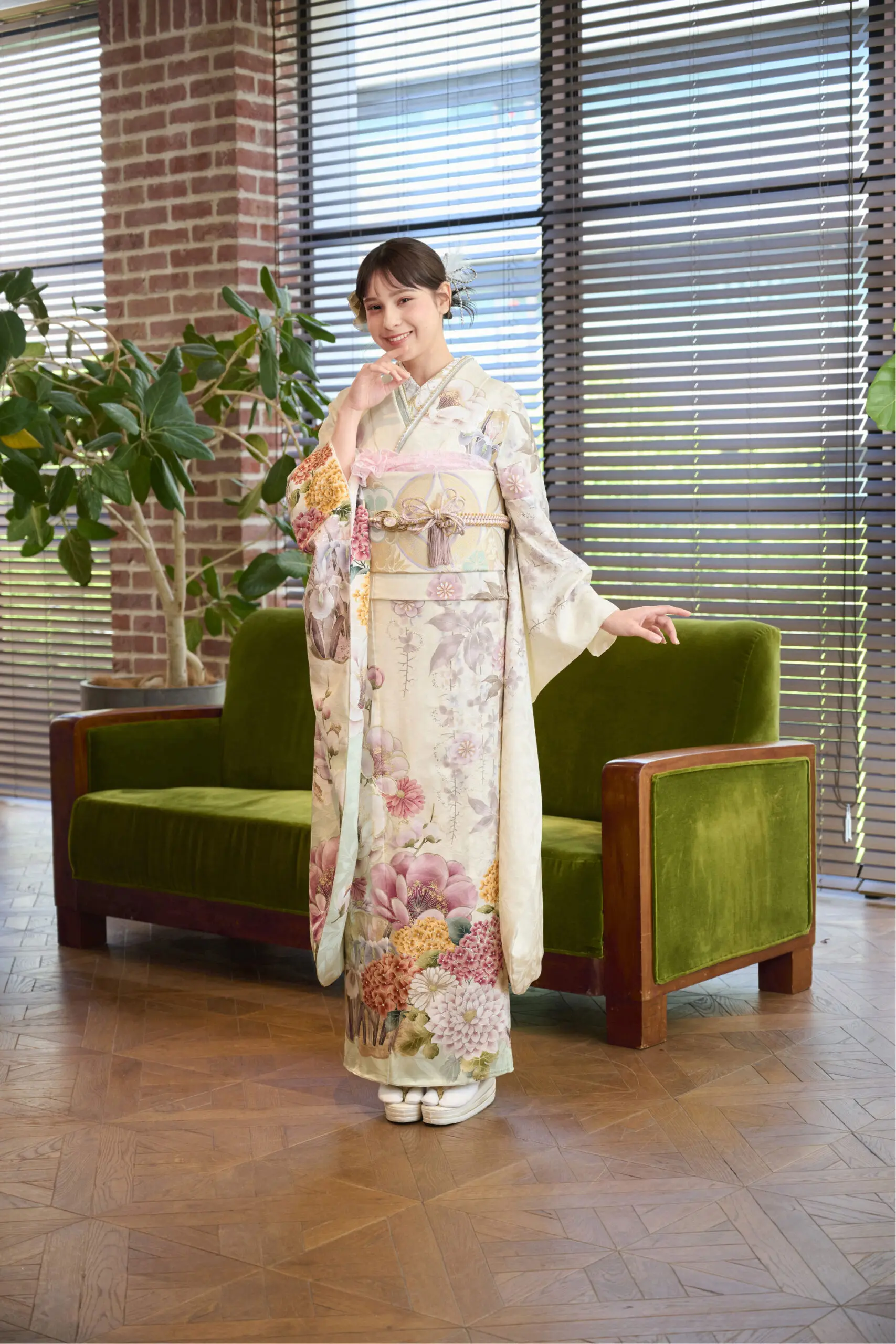 緑色のソファと大きな窓のそばで、M-417の花柄の着物に帯を締めて微笑む女性。鉢植えのそばに立ち、片手をあごに添えている。.