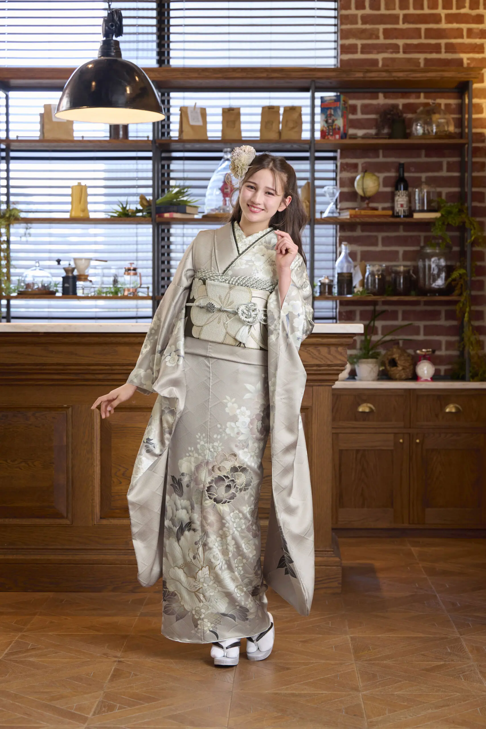 淡い色の花柄の着物に白い伝統的な履物を合わせたM-409を着て、室内でポーズをとる女性。木製の棚やキャビネット、装飾品に囲まれた背景で、片腕を少し上げて微笑んでいる。.