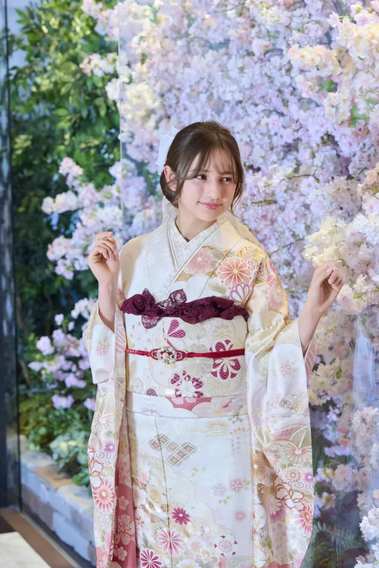 ピンクと白の桜の背景の前でポーズをとる若い女性。M-408：伝統的な淡い色の花模様の着物を着ている。両手を少し上げて立ち、横を見つめている。.